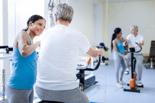 Friendly smile. Positive helpful professional worker of a rehabilitation center looking at her patient and smiling while helping him with the exercise bike
