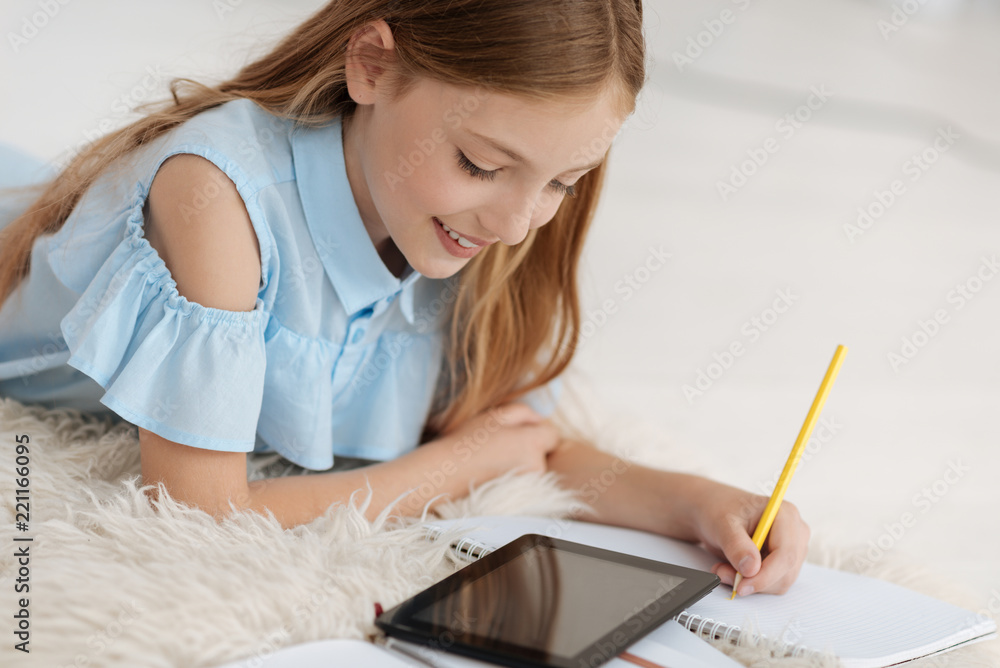 Be attentive. Charming little female making notes while looking at tablet and lying on the fluffy carpet Be attentive. Charming little female making notes while looking at tablet and lying on the fluffy carpet