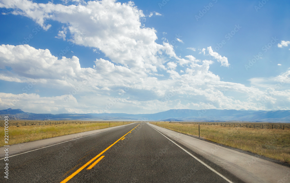 A straight highway cutting between pasture land disappearing into the distant blue mountains in a cloudy afternoon Wyoming landscape A straight highway cutting between pasture land disappearing into the distant blue mountains in a cloudy afternoon Wyoming landscape