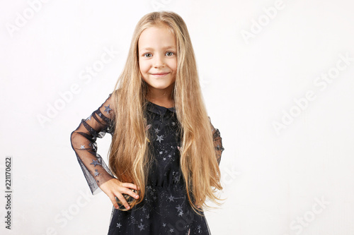 Little blonde girl with long golden hair wearing black dress with stars & planets, dancing, smling & having fun, white wall background. Five years old blonde female child posing. Copy space, close up.