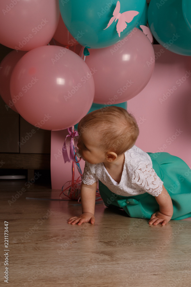 Obraz premium a little girl is sitting on the floor, on a pink background, near the balloons, with a pacifier. celebration. birthday, one year old. Little Girl with soother.
