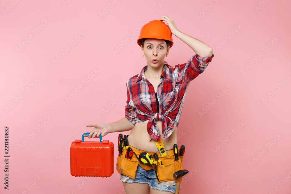 Strong young handyman woman in orange helmet, plaid shirt, denim shorts, kit tools belt full of instruments, toolbox isolated on pink background. Female in male work. Renovation and occupation concept Strong young handyman woman in orange helmet, plaid shirt, denim shorts, kit tools belt full of instruments, toolbox isolated on pink background. Female in male work. Renovation and occupation concept