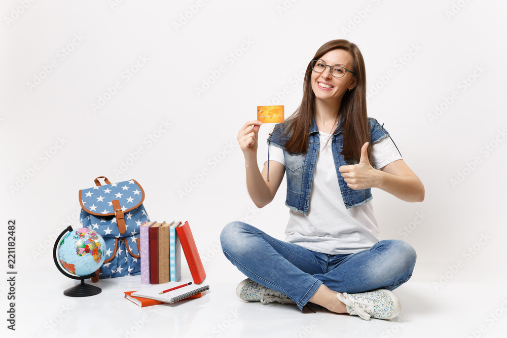 Young pretty smiling woman student in glasses holding credit card showing thumb up sitting near globe backpack, school books isolated on white background. Education in high school university college. Young pretty smiling woman student in glasses holding credit card showing thumb up sitting near globe backpack, school books isolated on white background. Education in high school university college.