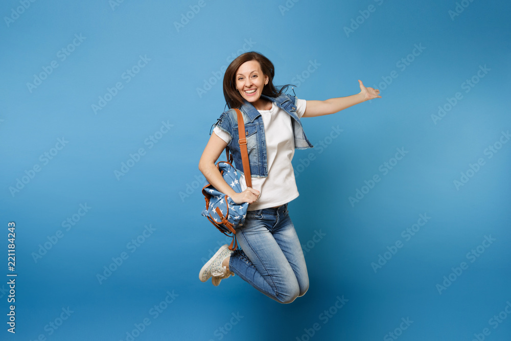 Full length portrait of young happy joyful woman student in denim clothes with backpack jumping and spreading hands isolated on blue background. Education in college. Copy space for advertisement. Full length portrait of young happy joyful woman student in denim clothes with backpack jumping and spreading hands isolated on blue background. Education in college. Copy space for advertisement.