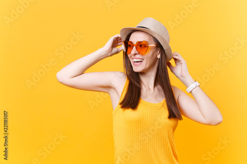 Portrait of excited smiling young woman in straw summer hat, orange glasses put hands on head, look aside isolated on yellow background. People sincere emotions, lifestyle concept. Advertising area.