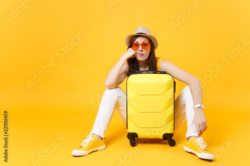 Bored tourist woman in summer casual clothes hat sit with suitcase, wait for plane isolated on yellow orange background. Girl traveling abroad to travel on weekends getaway. Air flight journey concept