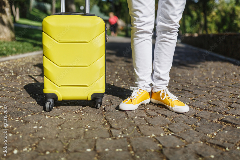 Fototapeta premium Cropped shot legs in yellow sneakers, tourist woman in casual clothes with suitcase standing, walking in city outdoor. Girl traveling abroad to travel on weekends getaway. Tourism lifestyle concept.