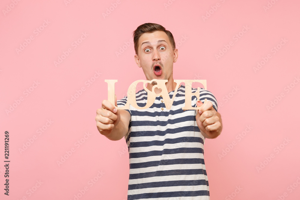 Portrait of emotional young man in striped t-shirt hold wooden word Love isolated on trending pastel pink background. People sincere emotions lifestyle holiday concept. Advertising area for copy space Portrait of emotional young man in striped t-shirt hold wooden word Love isolated on trending pastel pink background. People sincere emotions lifestyle holiday concept. Advertising area for copy space