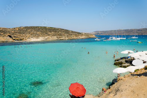 Crystal clear turqoise sea color on island Comino in famous Blue lagoon. Aerial beach scene with parasols against sun during summer holiday vacation on Malta. Blue sky, clear water with sandy bottom.