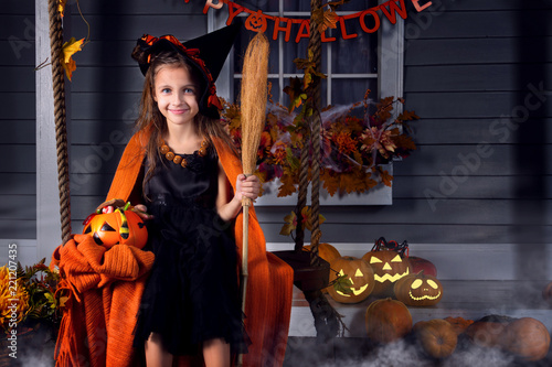 Funny girl child kid in halloween witch costume with black hat and orange pumpkins basket with spooky face for candies and sweets sitting on yard swing ready for celebration autumn holiday halloween.