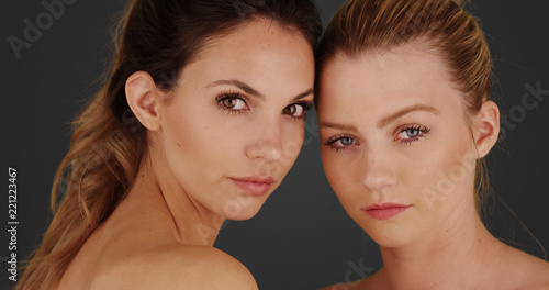 Headshot of young blonde and brunette with makeup on posing on gray background.