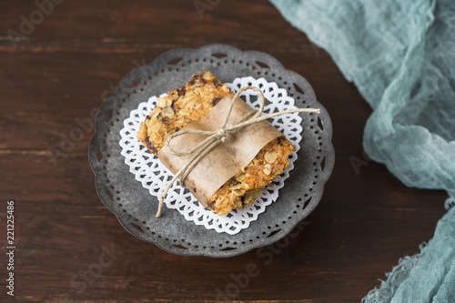 Homemade Chewy Granola Bars on a Tin Pedestal on a Wood Table; Granola Bar Wrapped in Parchment Paper and String; White Heart-Shaped Doily on Pedestal