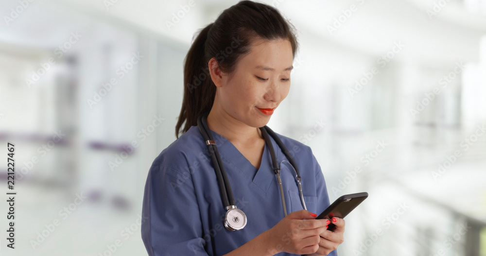 Happy young Asian doctor in scrubs texting with smartphone inside empty hospital Happy young Asian doctor in scrubs texting with smartphone inside empty hospital