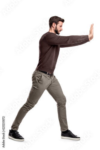 A bearded man in casual clothes tries to push a heavy object with both arms with one leg put in front for balance. photo