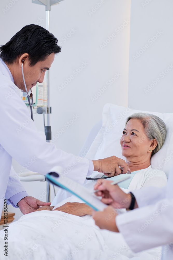 Doctor checking heartbeat of senior female patient and nurse taking notes Doctor checking heartbeat of senior female patient and nurse taking notes