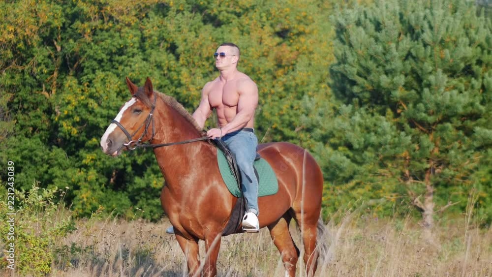 Athletic strong man with naked torso in sunglasses sitting on a horse in a field near the forest Athletic strong man with naked torso in sunglasses sitting on a horse in a field near the forest