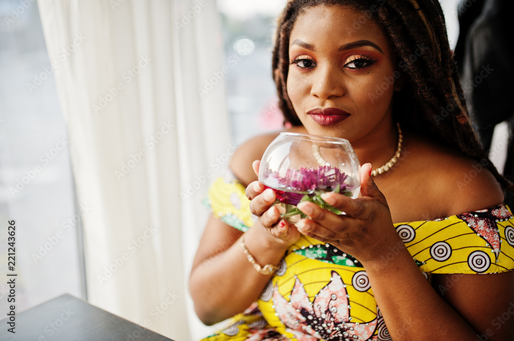 Cute small height african american girl with dreadlocks, wear at coloured yellow dress, sitting at cafe with flower at glass. Cute small height african american girl with dreadlocks, wear at coloured yellow dress, sitting at cafe with flower at glass.