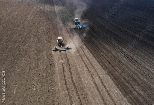 Aerial view of two blue tractors plows the earth in field on a summer day against a black earth background. Agriculture. Two tractors travel one after another along the black field