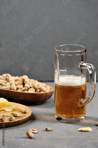 A mug of light beer. As a snack for beer pistachios in a bowl and potato chips on a taella. Gray wooden background. Close-up. Vertical orientation of the frame.