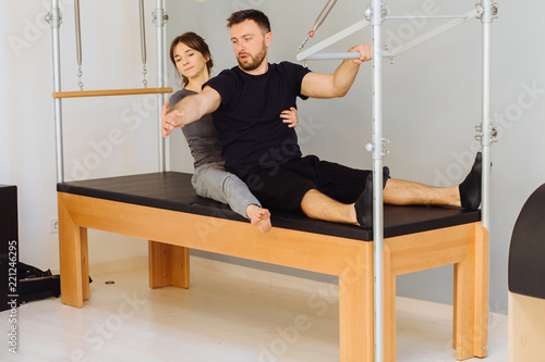 Young sporty female instructor of pilates helping beard man workout in cadillac bed. Two people working in pilates studio, woman assistant supporting and correcting male patient beginner.