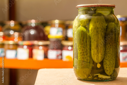 Homemade glass jar with pickled cucumbers in the cellar on the background of shelves with canned vegetables and fruits. Closeup, selective focus
