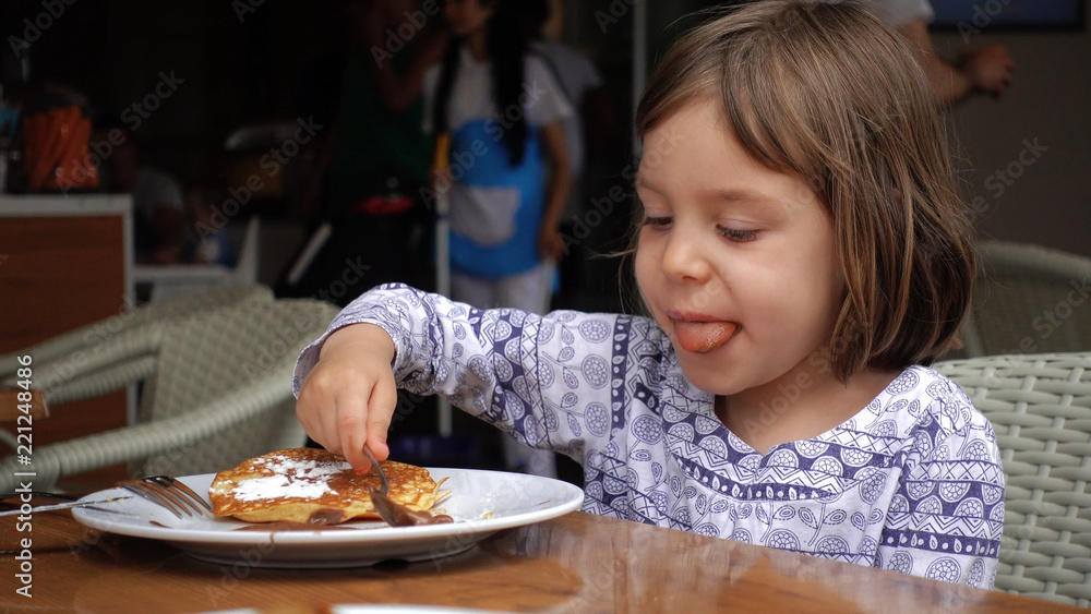Girl eats pancakes, poured with chocolate and sprinkled with powdered sugar.