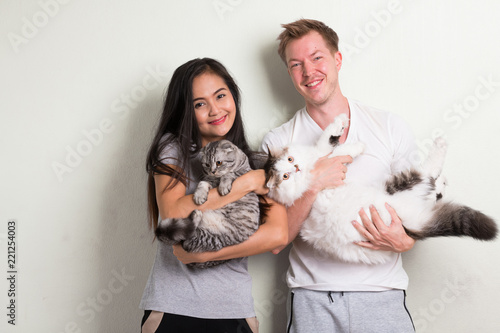 Young happy multi-ethnic couple holding two cats together against white background