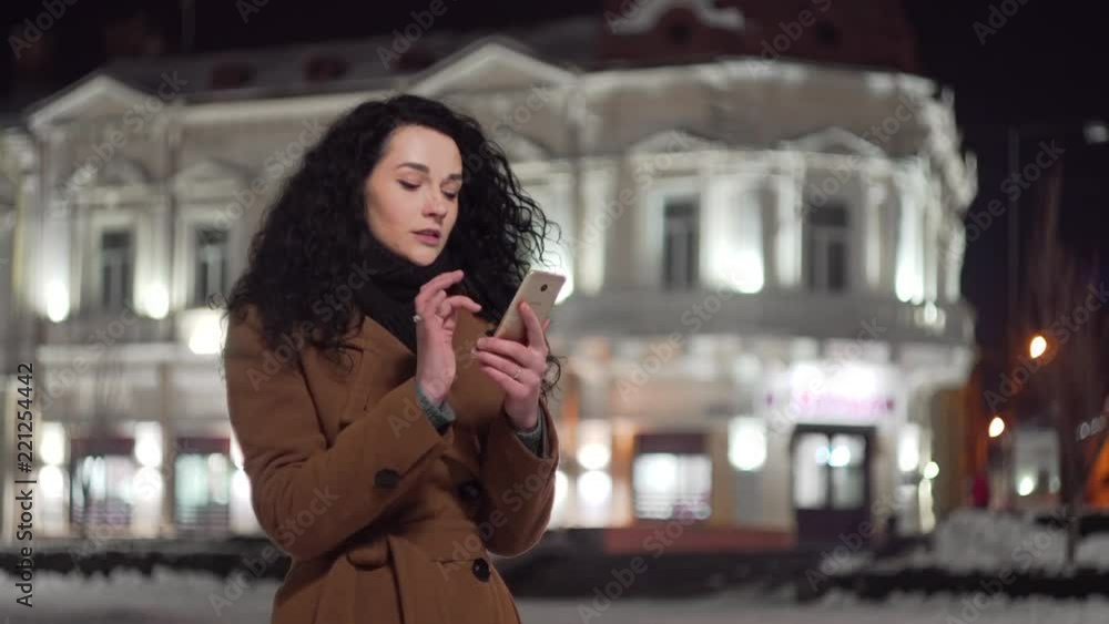 Portrait of young beautiful woman using smart phone hand hold outdoor in the city night, smiling, face illuminated screenlight - social network, technology, comunication concept Portrait of young beautiful woman using smart phone hand hold outdoor in the city night, smiling, face illuminated screenlight - social network, technology, comunication concept