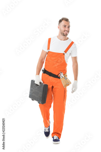 handsome auto mechanic in orange uniform holding toolbox and looking away isolated on white
