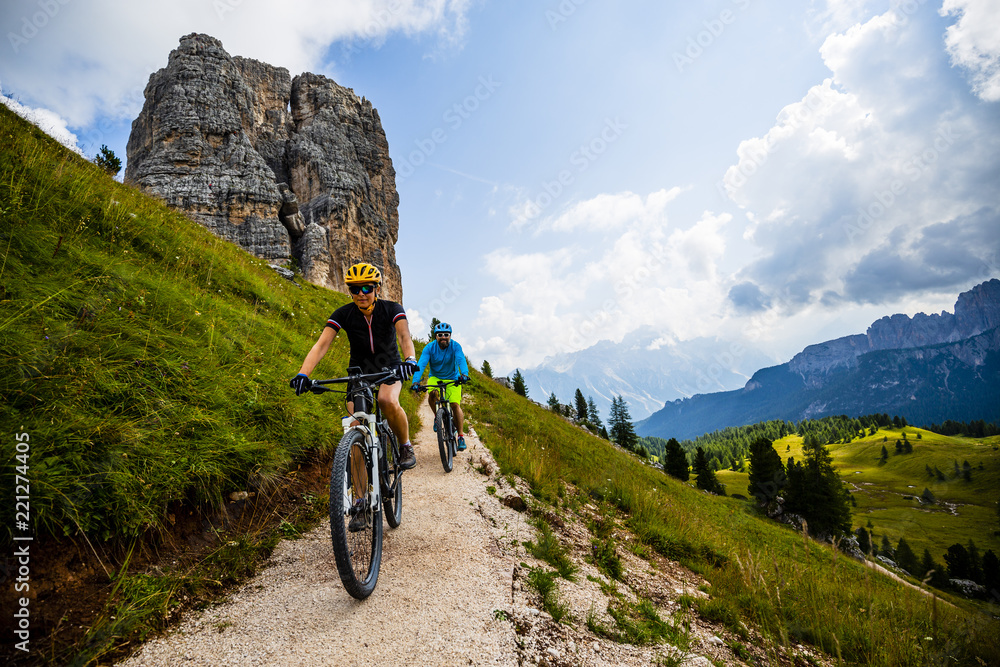 Couple cycling in Cortina d'Ampezzo, stunning Cinque Torri and Tofana in background. Woman and man riding MTB trail. South Tyrol province of Italy, Dolomites.