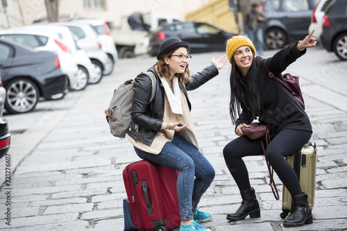 Due amiche in viaggio si siedono sopra le valige e indicano qualcosa photo