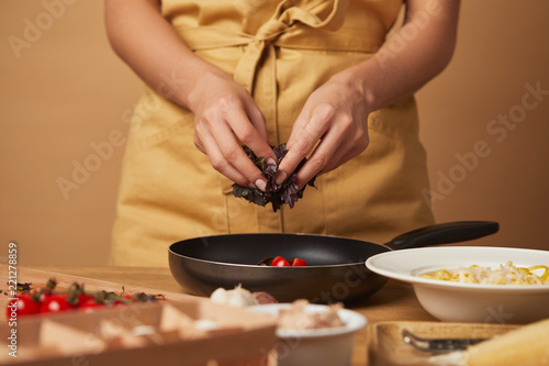 cropped shot of woman putting basil into frying pan while cooking pasta