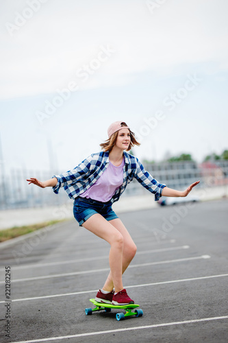 A beautiful smiling blond girl wearing checkered shirt, cap and denim shorts is longboarding while stretching out her hands. Sport and cool style.