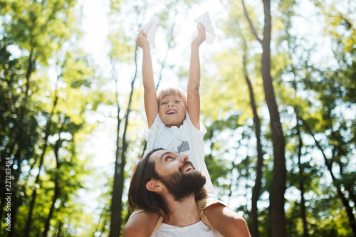 Handsome father and his little son walking in the forest. Boy is sitting on the shoulders of his father and keeps a paper airplane in his hand.