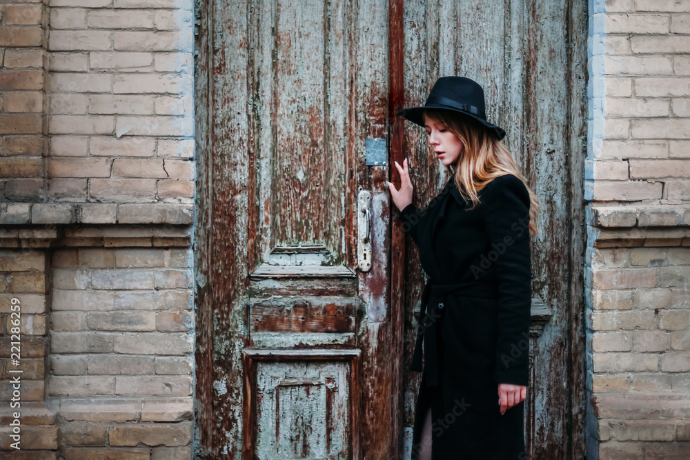 Blonde beautiful girl with long hair, in black coat in hat, stands on the background of vintage antique old wooden door old brick house. For concept design. Blonde beautiful girl with long hair, in black coat in hat, stands on the background of vintage antique old wooden door old brick house. For concept design.