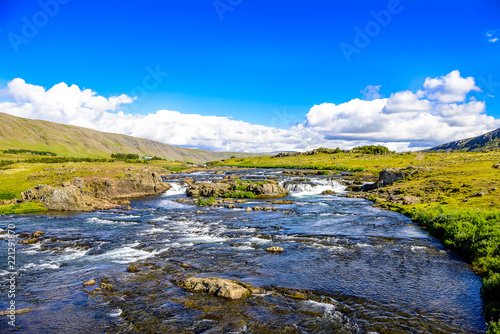 Beautiful Iceland Waterfall.Fast running river. Rugged volcanic rock cliffs against blue skies with low clouds. Green moss and grass covered rock formation.