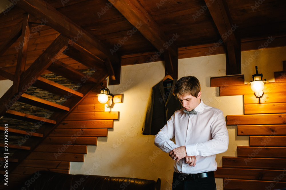 Fototapeta premium A man in a shirt with a bow tie , wears a metallic silver cufflinks stud on his arm. On the background of a brown jacket hanging on a wall. Hand's groom. Wedding day.