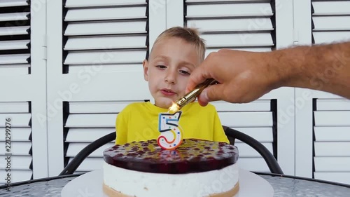 Little boy sitting at table and blowing candles on birthday cake. Cute happy boy celebrating its fifth birthday, in front of him delicious cake with candle in the form of 5 photo