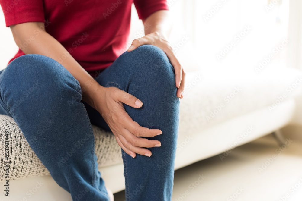 Closeup young woman sitting on sofa and feeling knee pain and she massage her knee at home. Healthcare and medical concept.