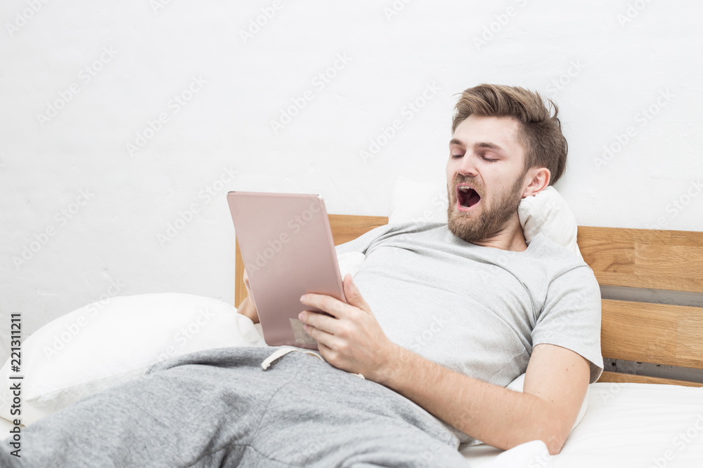 Caucasian young man holding tablet and yawning want to sleep feeling so tried on bed in bedroom Caucasian young man holding tablet and yawning want to sleep feeling so tried on bed in bedroom