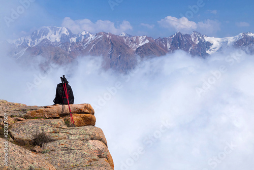 Black backpack and red trekking poles over a steep mountain slope, snowy mountains over clouds on the background