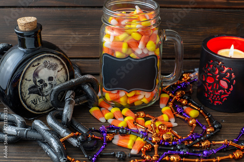 Halloween background frame with a jar of candy corn on a wooden table surrounded by a dark chain and next to a vial of poison and day of the dead candle with a black lable that you can personalize.