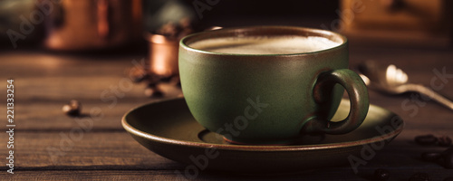 Green cup of coffee with beans and coffee mill on old wooden background with copy space. Retro style toned dark photography. Banner.