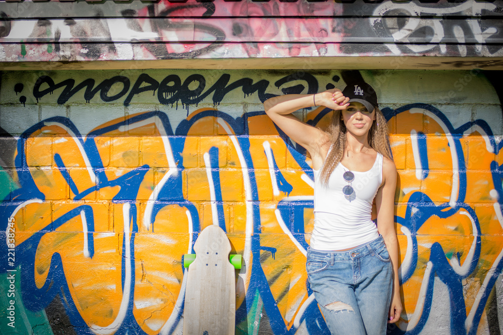 Fototapeta premium Portrait of young beautiful woman wearing white tank shirt and blue jeans and black hat on brick wall with graffiti background