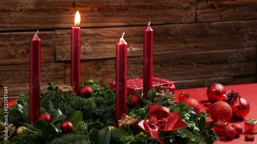 One burning red candle on a traditional advent wreath of green fir twigs and mistletoes with festive decoration in front of a rustic wooden wall, close-up real time shot with copy space, nobody, 30s