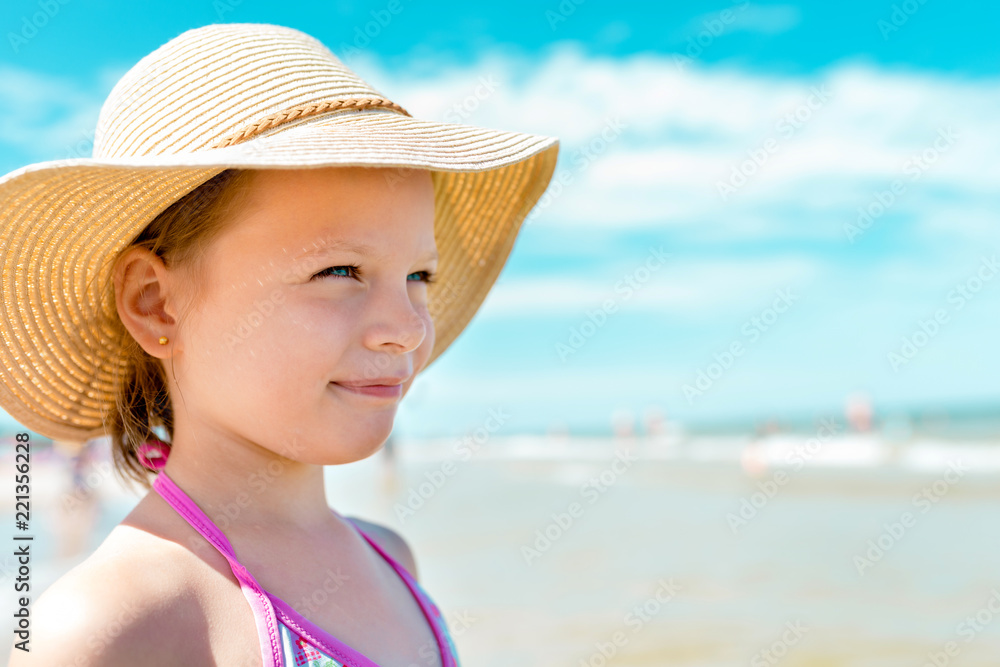 Adorable little girl in a straw hat on the beach  during summer vacation, cloudy sky in the background