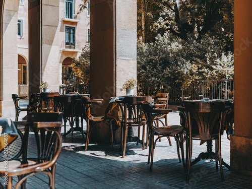 Cozy outdoor cafe with empty wooden tables covered with a tablecloth with metal flower pots and ashtries on it  chairs near  exterior of an empty street bar on the daytime waiting for its visitors