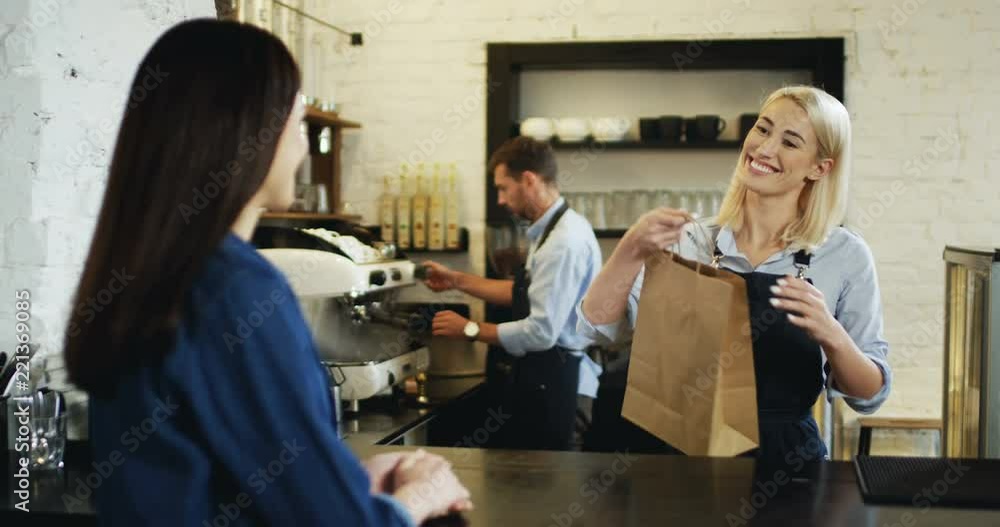 Caucasian woman buying something at the bar in the cafe and pretty blonde waitress giving her a cartoon bag, then she going away. Waiter making coffee at the background. Caucasian woman buying something at the bar in the cafe and pretty blonde waitress giving her a cartoon bag, then she going away. Waiter making coffee at the background.