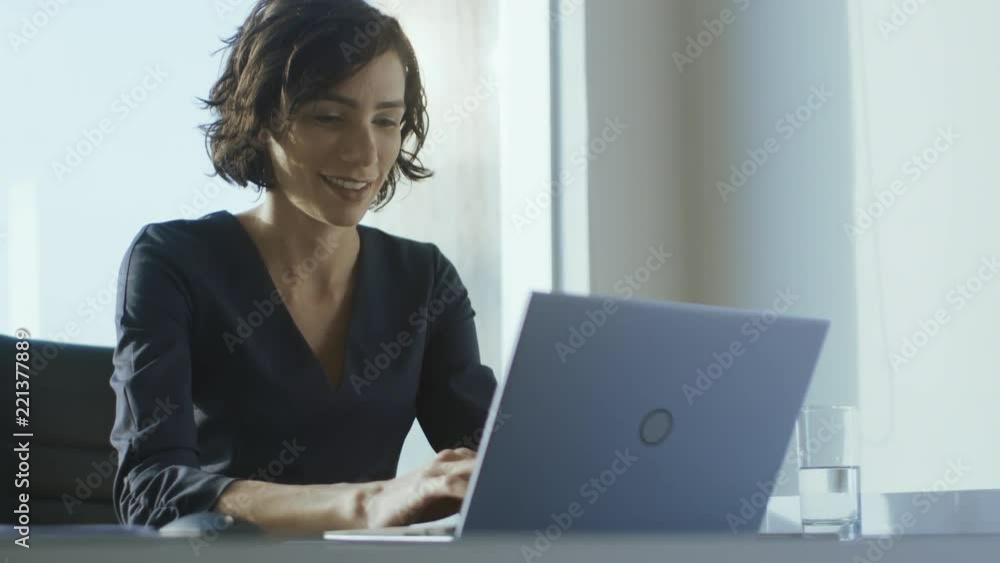 Confident Female Executive Works on a Laptop Sitting at Her Desk in Modern Office with Big City View. Smiling Successful Businesswoman Uses Laptop. Confident Female Executive Works on a Laptop Sitting at Her Desk in Modern Office with Big City View. Smiling Successful Businesswoman Uses Laptop.
