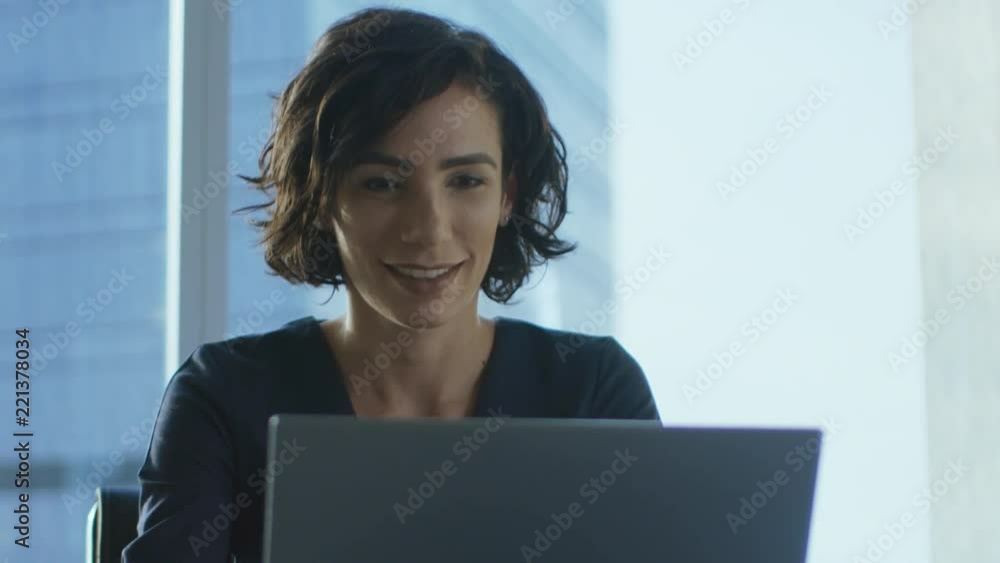 Confident Female Executive Works on a Laptop Sitting at Her Desk in Modern Office with Big City View. Smiling Successful Businesswoman Uses Laptop. Confident Female Executive Works on a Laptop Sitting at Her Desk in Modern Office with Big City View. Smiling Successful Businesswoman Uses Laptop.